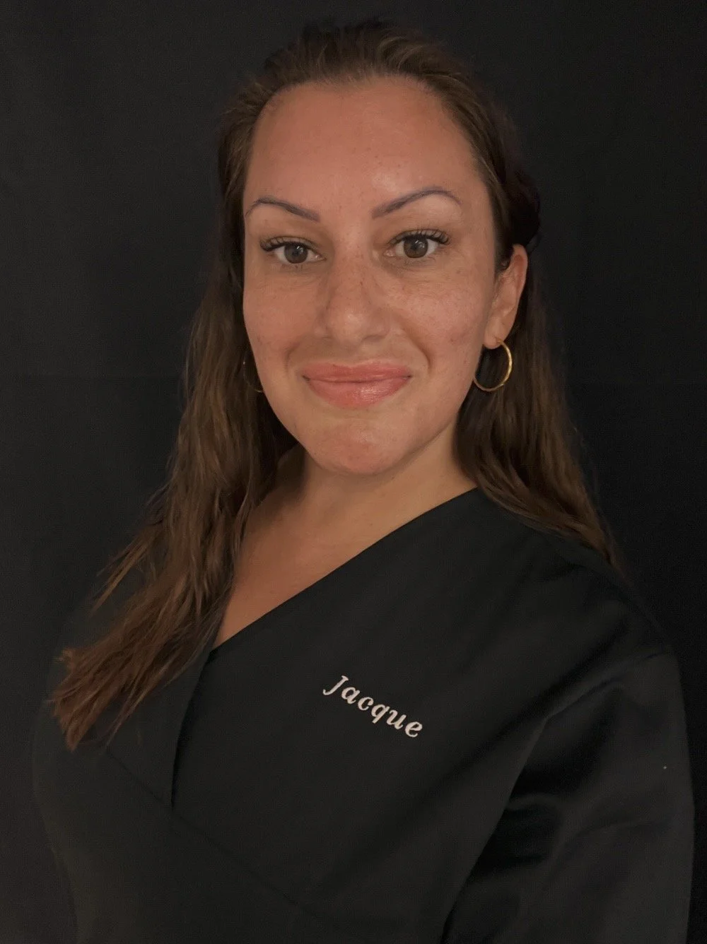 A woman with long, wavy brown hair, wearing a black top with the name 'Jacque' embroidered on it, gold hoop earrings, and natural makeup, smiling against a black background.