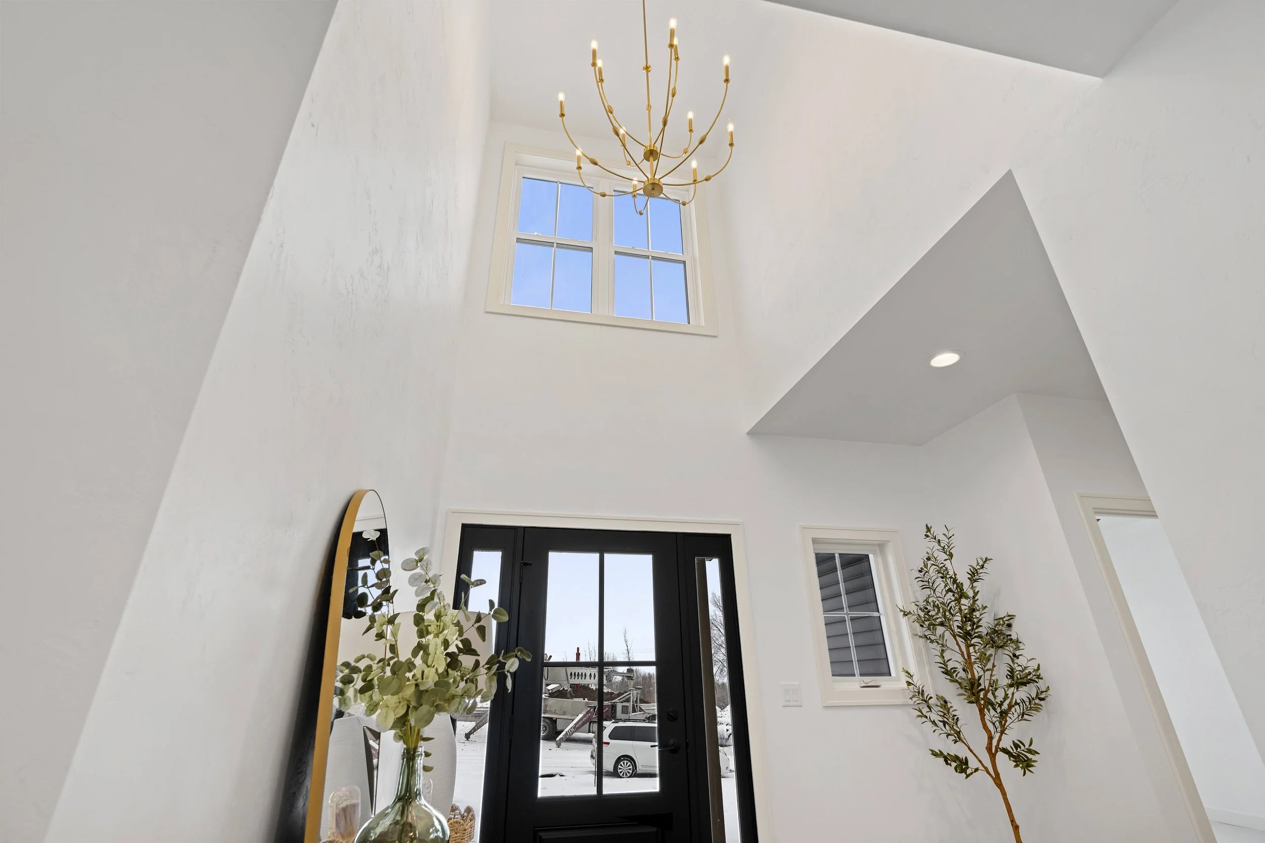 Modern entryway with white walls, black front door with glass panes, a tall window above the door, a hanging chandelier, a large mirror, a houseplant, and snow outside.