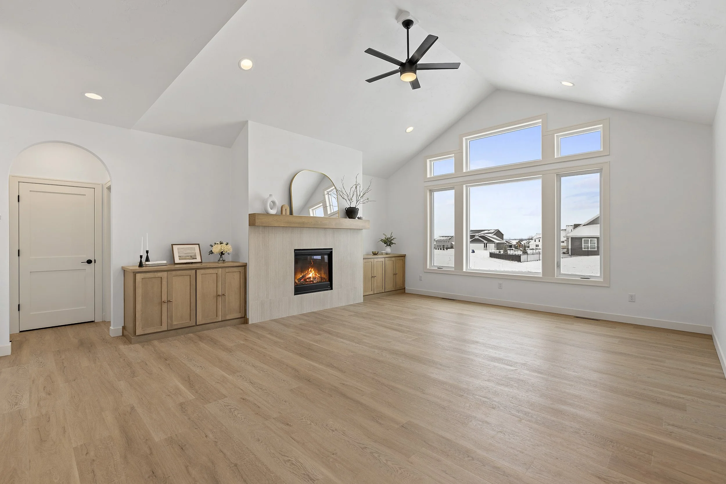 Empty living room with white walls, large window view of snowy neighborhood, wood flooring, fireplace, and ceiling fan.