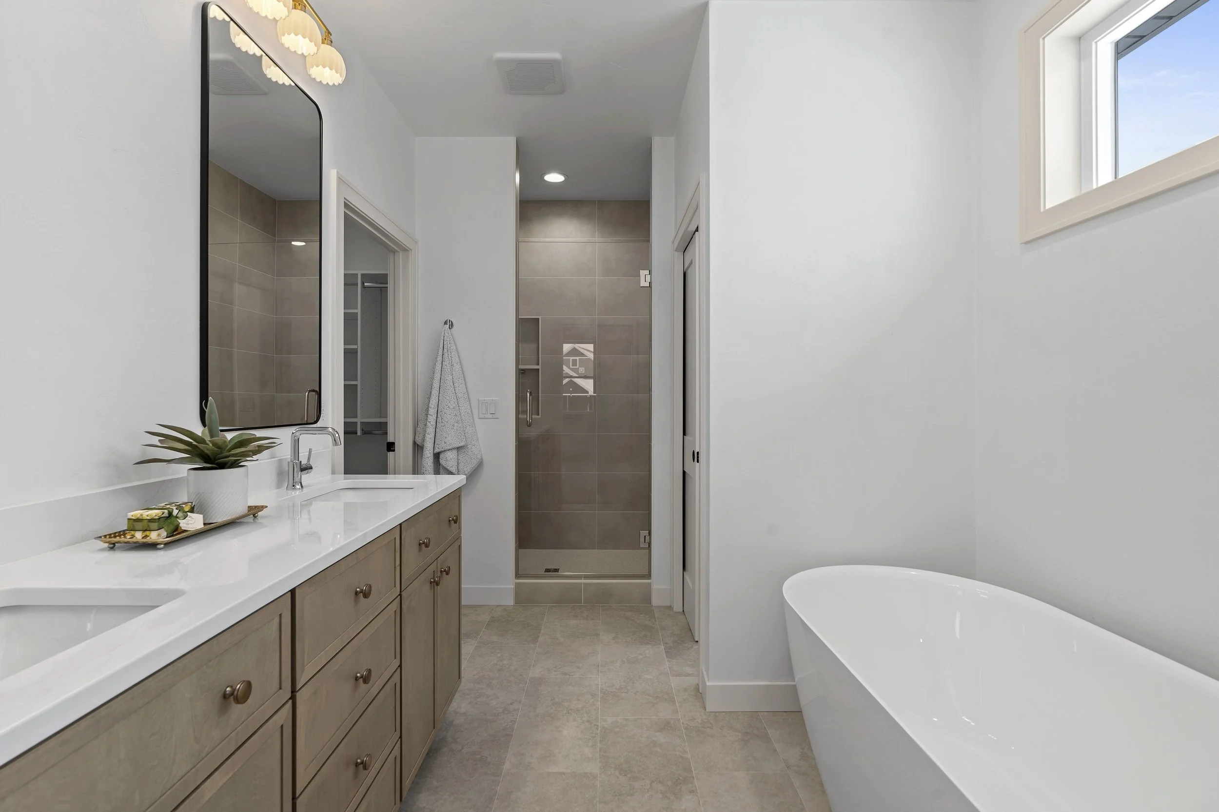 Modern bathroom with double vanity, large mirror, potted plant, walk-in shower, and freestanding bathtub, with neutral colors and natural light.