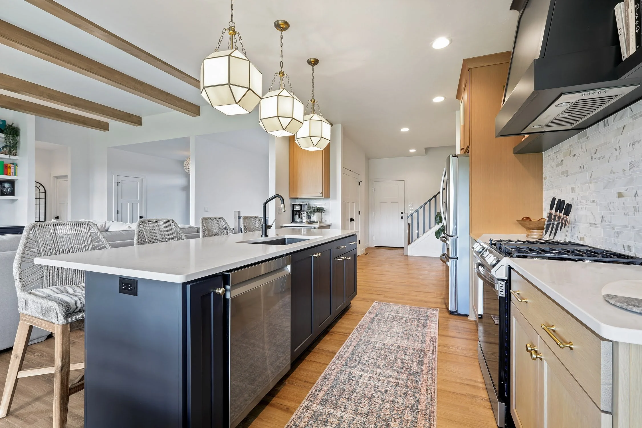 Modern kitchen with black island, white countertops, light wood cabinets, stainless steel appliances, a row of pendant lights, hardwood floors, and a small rug.