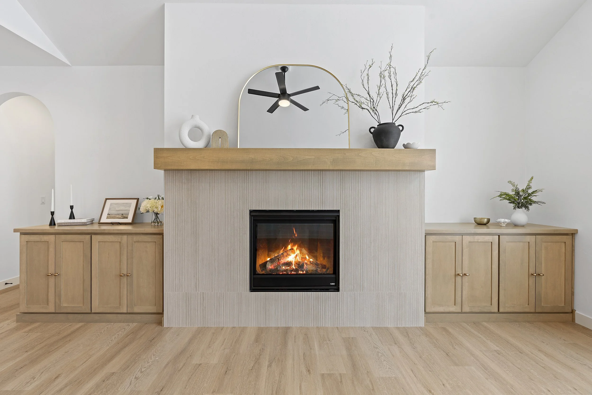Modern living room with a wood fireplace, wooden cabinets on each side, and decorative items like vases, picture frame, and plants on top of the cabinets and mantel. A ceiling fan is reflected in a mirror above the fireplace.