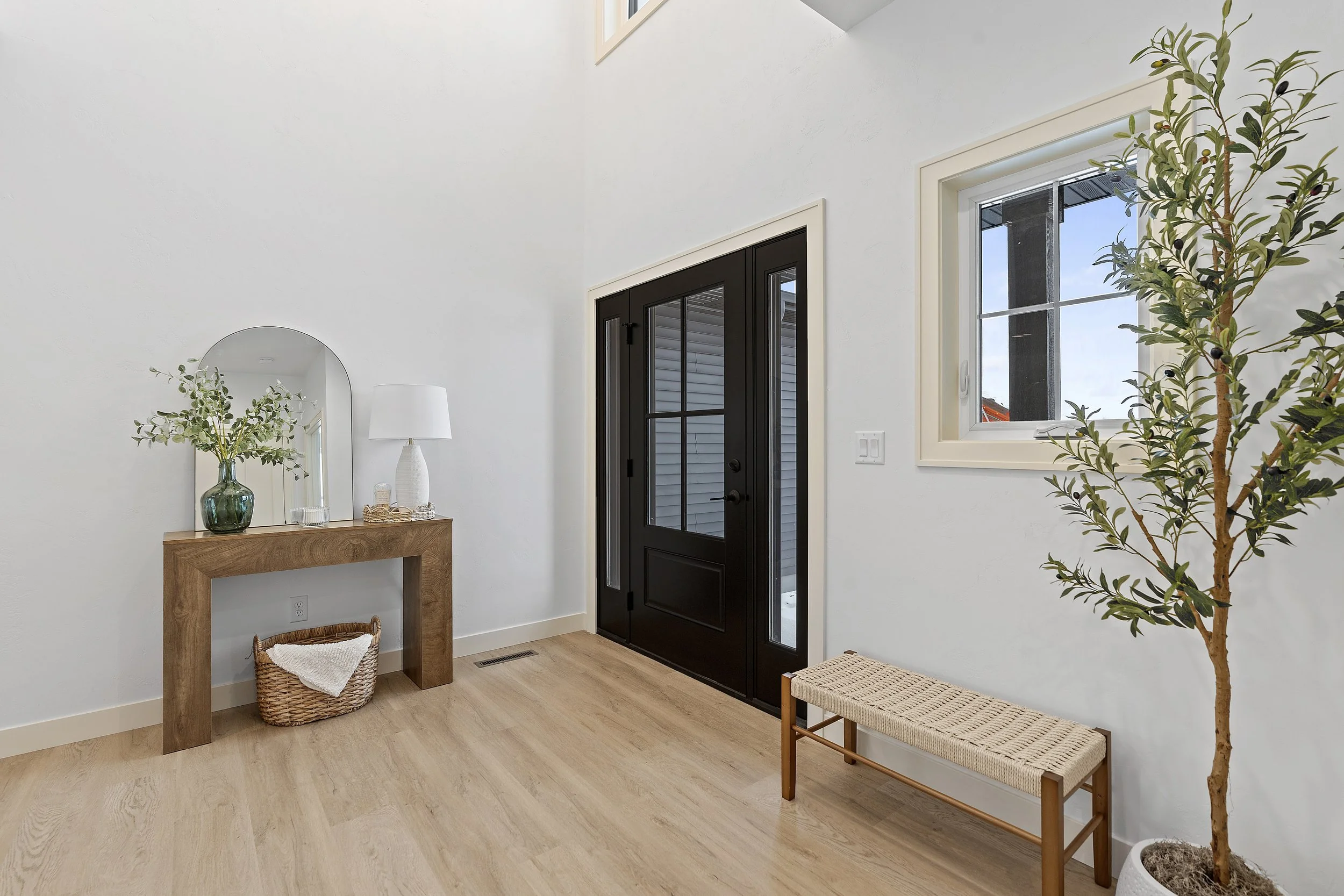 Entryway with black front door, window, wooden bench, and a small table with decor and a mirror, indoor plant, white walls, wood flooring.