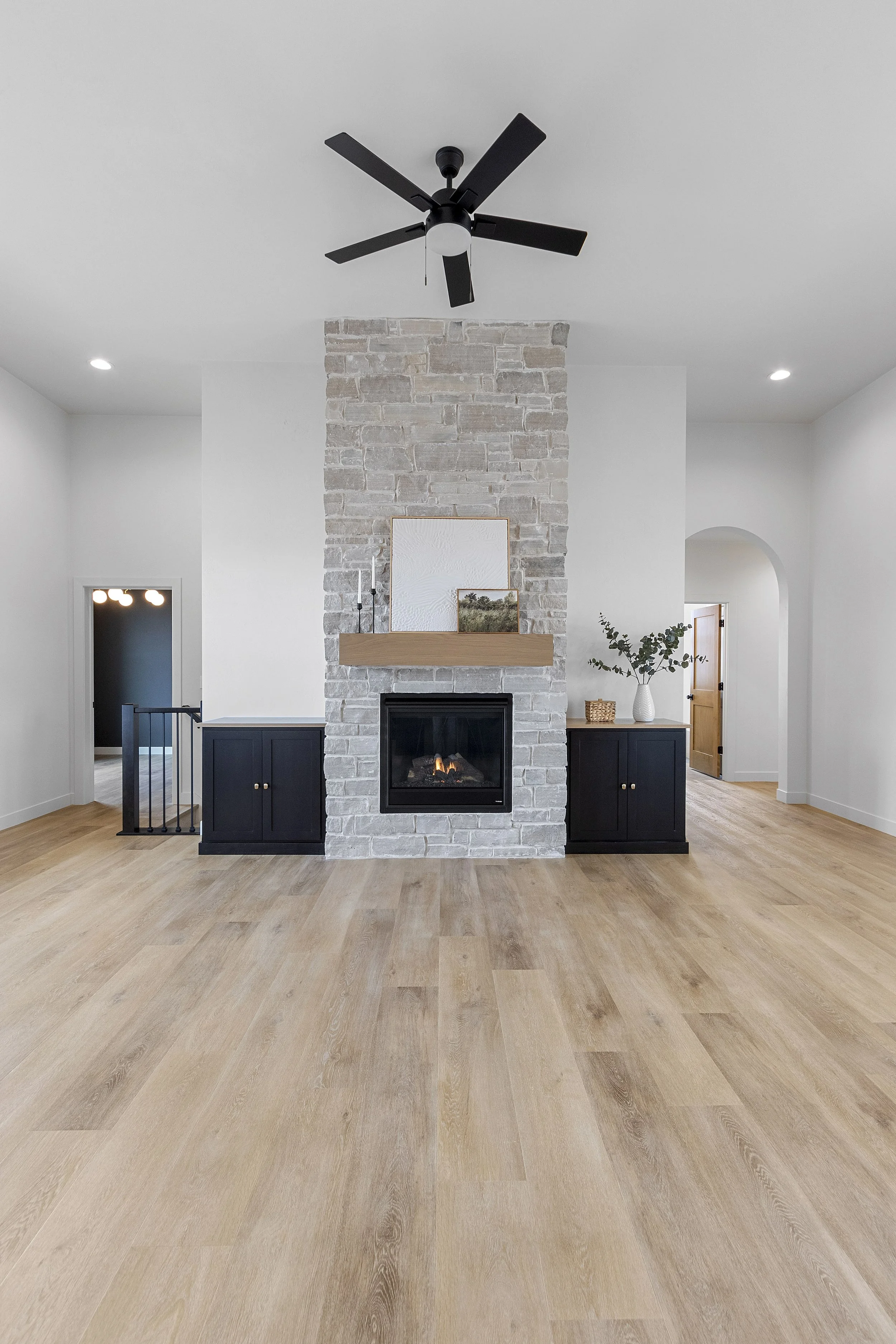 Living room with a brick fireplace, black cabinetry, hardwood flooring, a ceiling fan, and minimal decor.