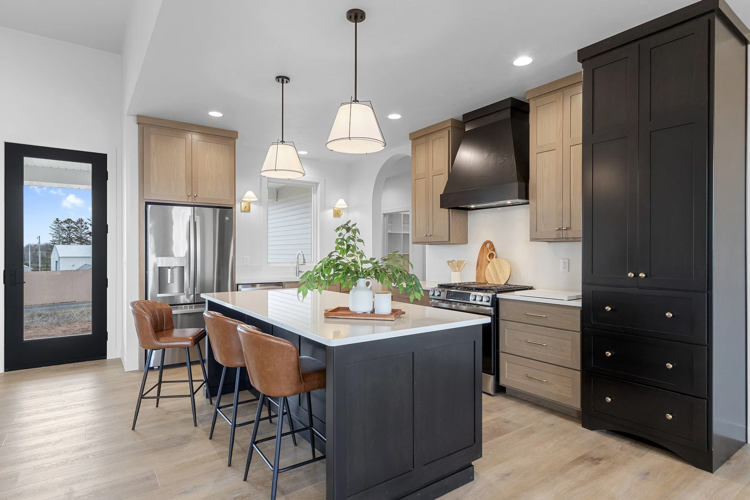 Modern kitchen with black island, wooden cabinets, stainless steel refrigerator, black range hood, and pendant lighting