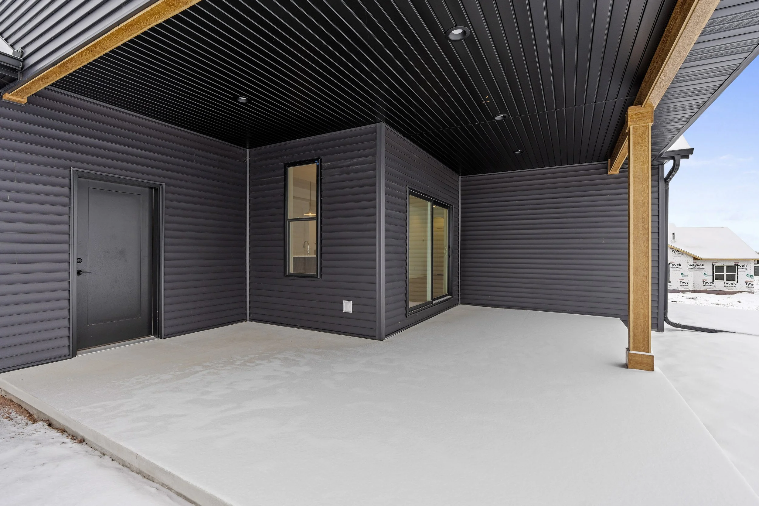 Covered outdoor patio with concrete floor, black siding, black door, and windows, with snow on the ground outside.