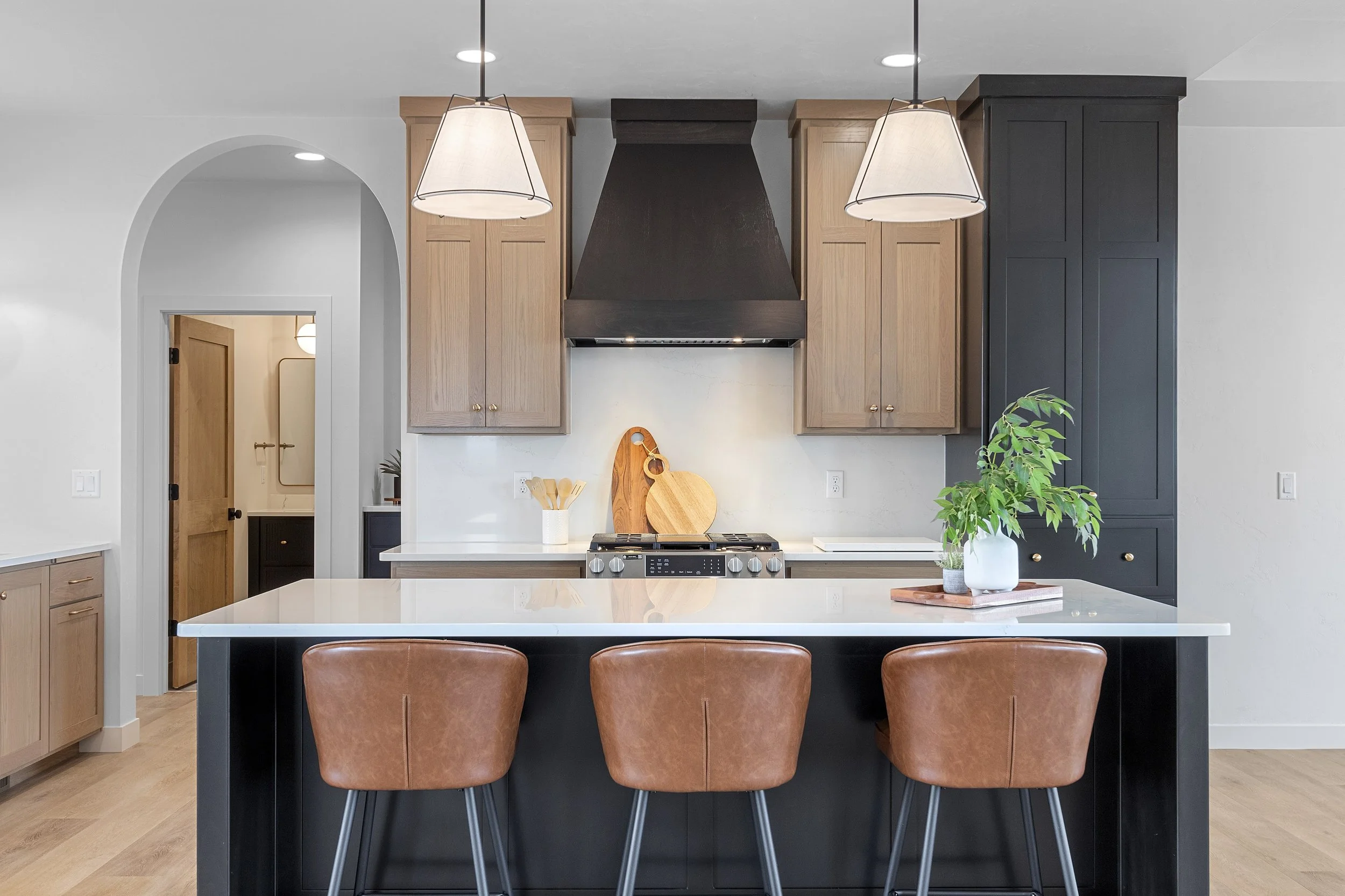 Modern kitchen with black island and three tan leather barstools, wooden cabinets, white countertops, hanging pendant lights, potted plant on the island, and cutting boards on the wall.