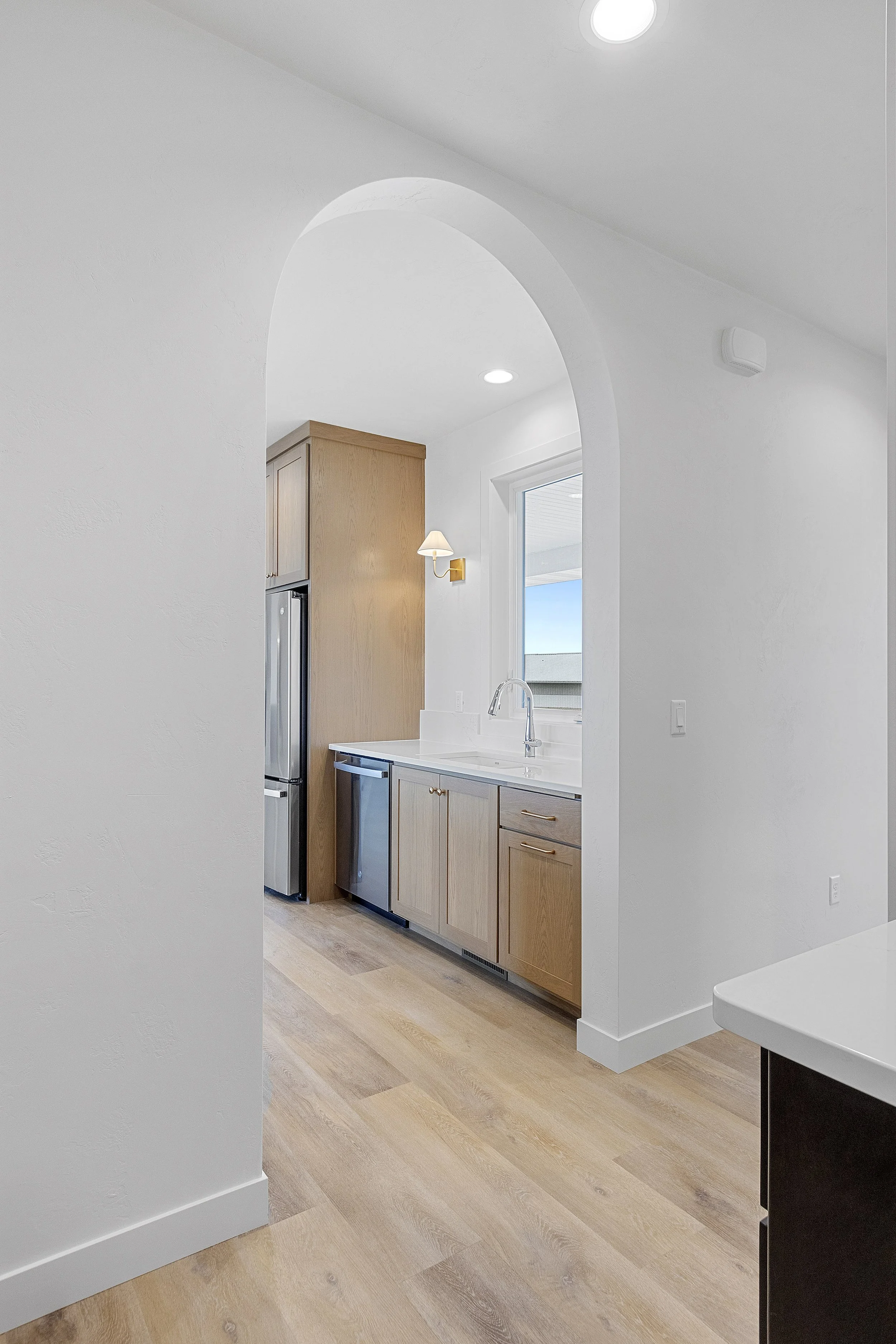 A view of a modern kitchen with light wood cabinets, a stainless steel refrigerator, a white countertop, a brass wall sconce, and natural light coming through a window, seen through an archway.