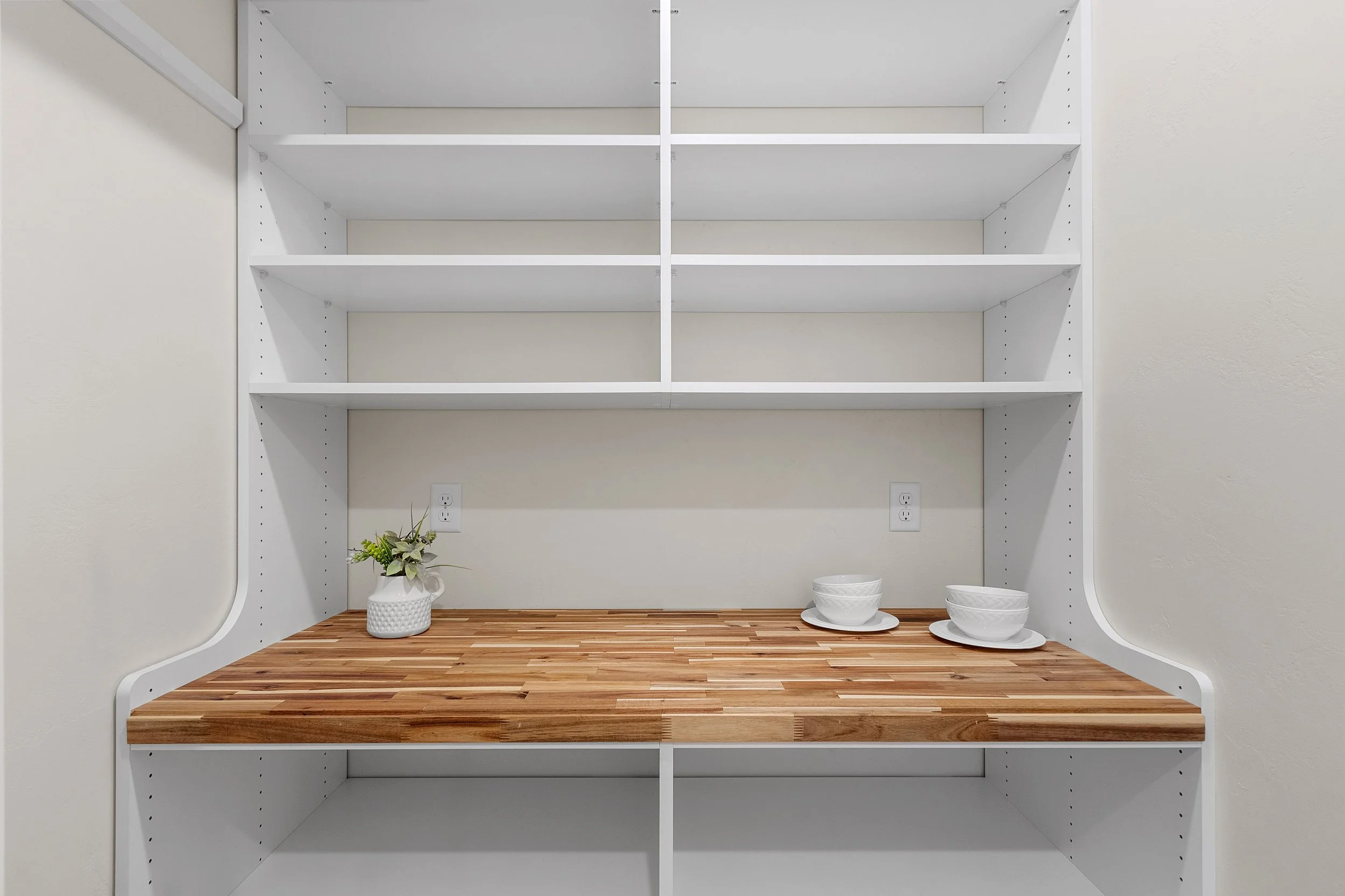 Empty white kitchen shelving unit with a wooden countertop, containing a white vase with green plants on the left and three white bowls on small white plates on the right, against a plain white wall with two electrical outlets.