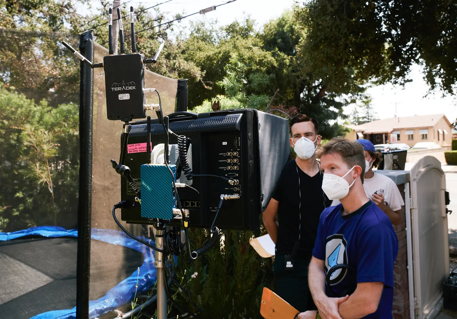 People wearing face masks on a film or video shoot outdoors, standing near a large monitor and equipment, with trees and houses in the background.