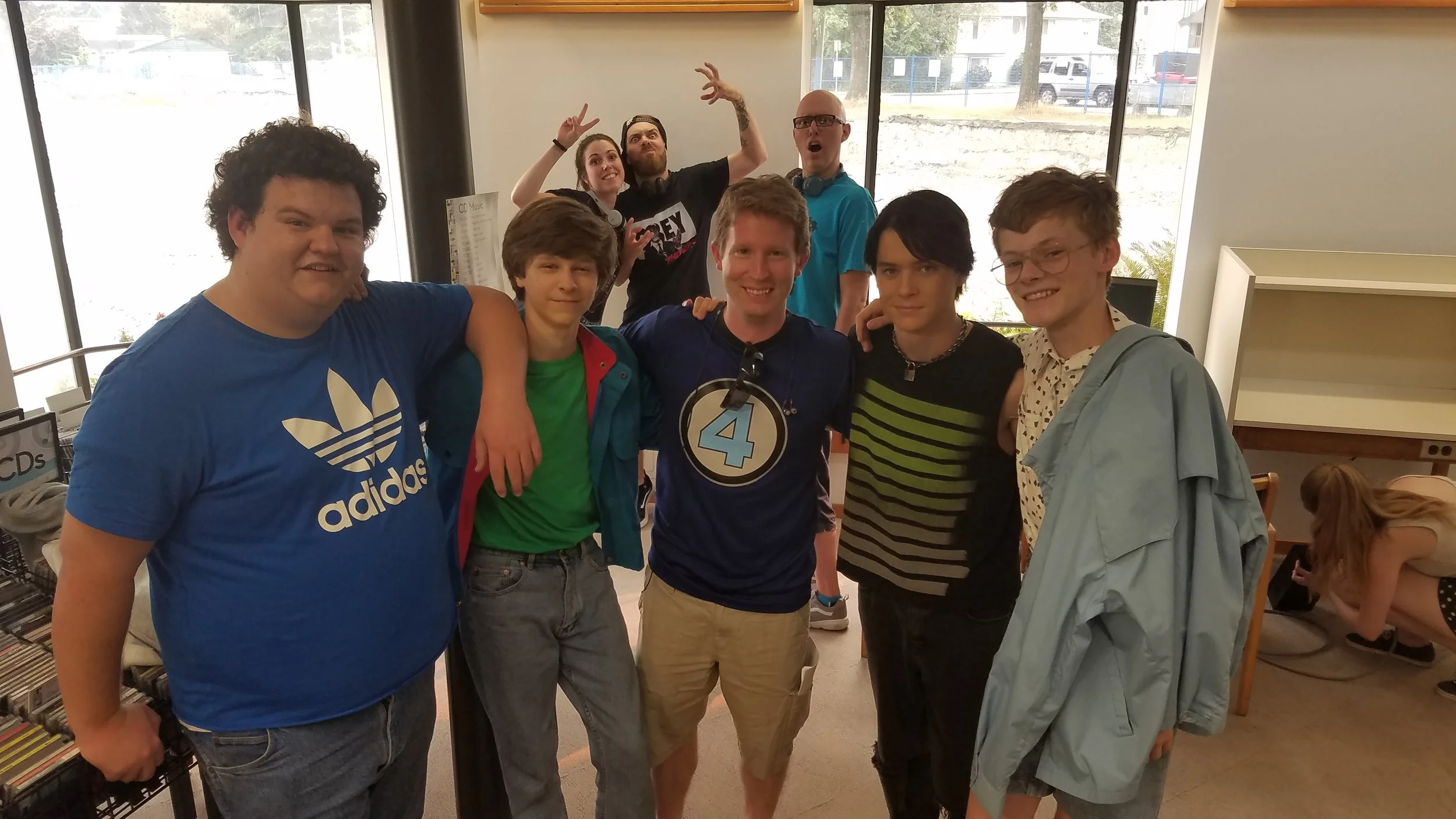 Group of seven young people posing together inside a building with large windows, some smiling and making peace signs, and one person in the background playfully making a face and raised hands.