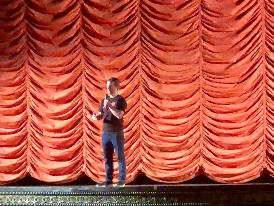 A man standing on stage with a microphone in front of a large red curtain.