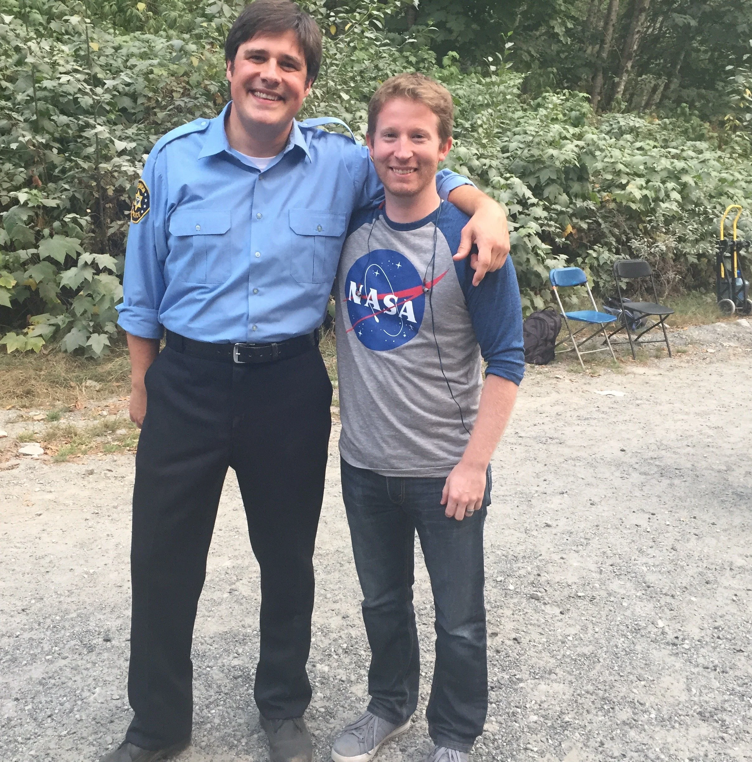 Two men standing outdoors with trees and chairs in the background. One is a large man in a blue uniform with a badge, and the other is a smaller man wearing a NASA t-shirt and jeans. They are smiling and have their arms around each other's shoulders.