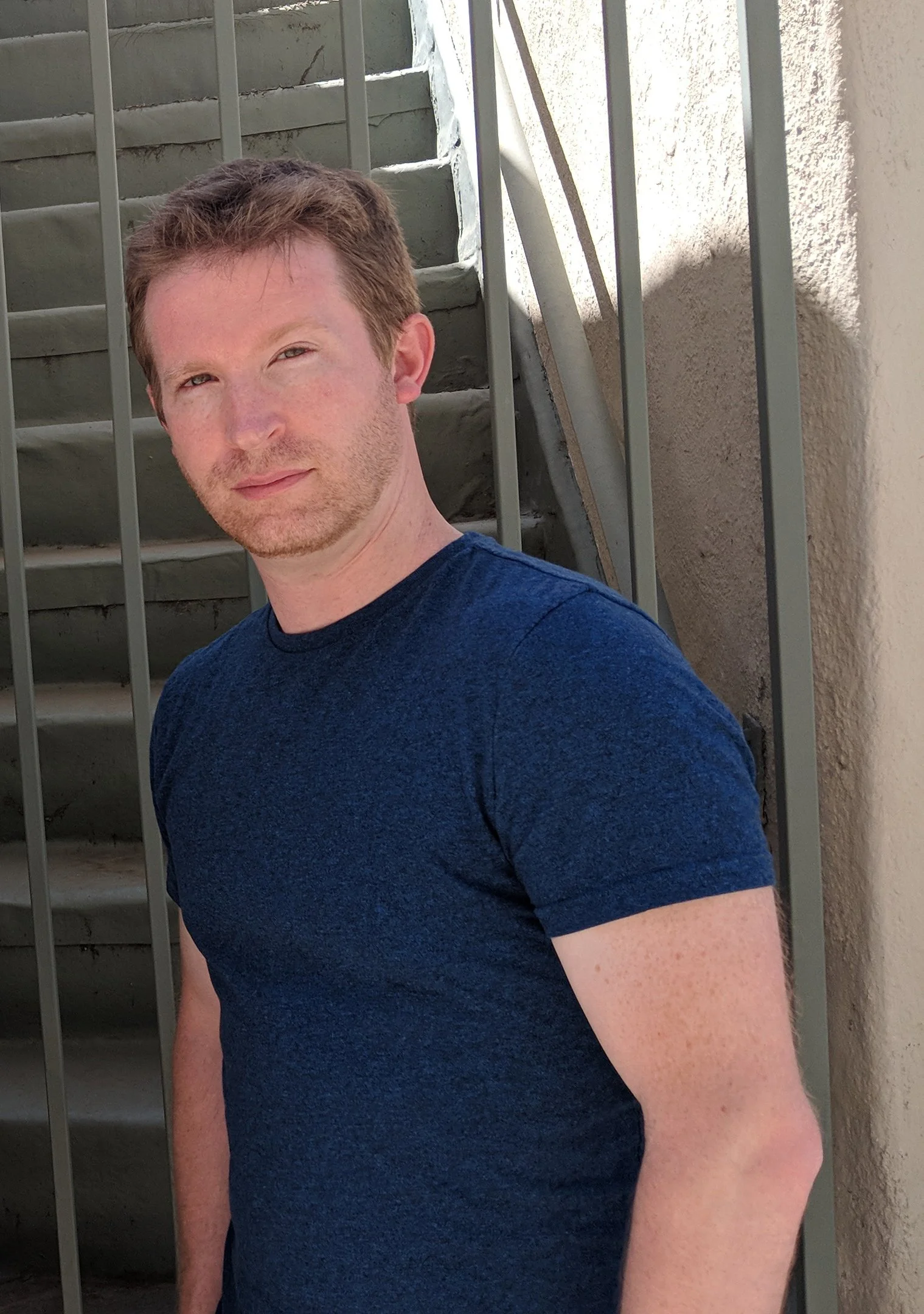 A man with light brown hair and a trim beard, wearing a navy blue T-shirt, standing outdoors near a staircase with metal railings, looking at the camera.
