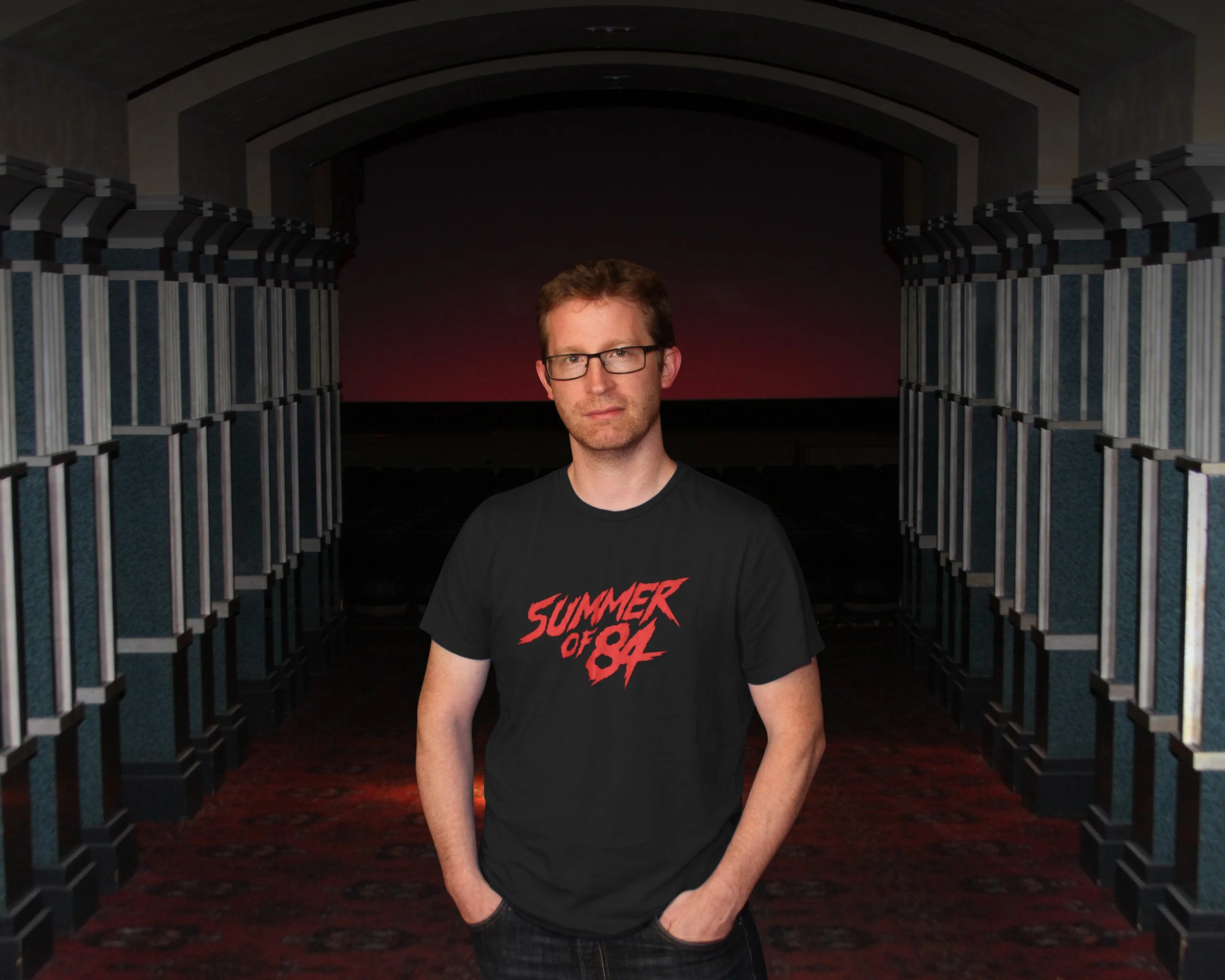 A man in glasses wearing a black t-shirt that says 'Summer of 84' stands in an empty movie theater lobby with rows of blue chairs and a red-lit screen in the background.