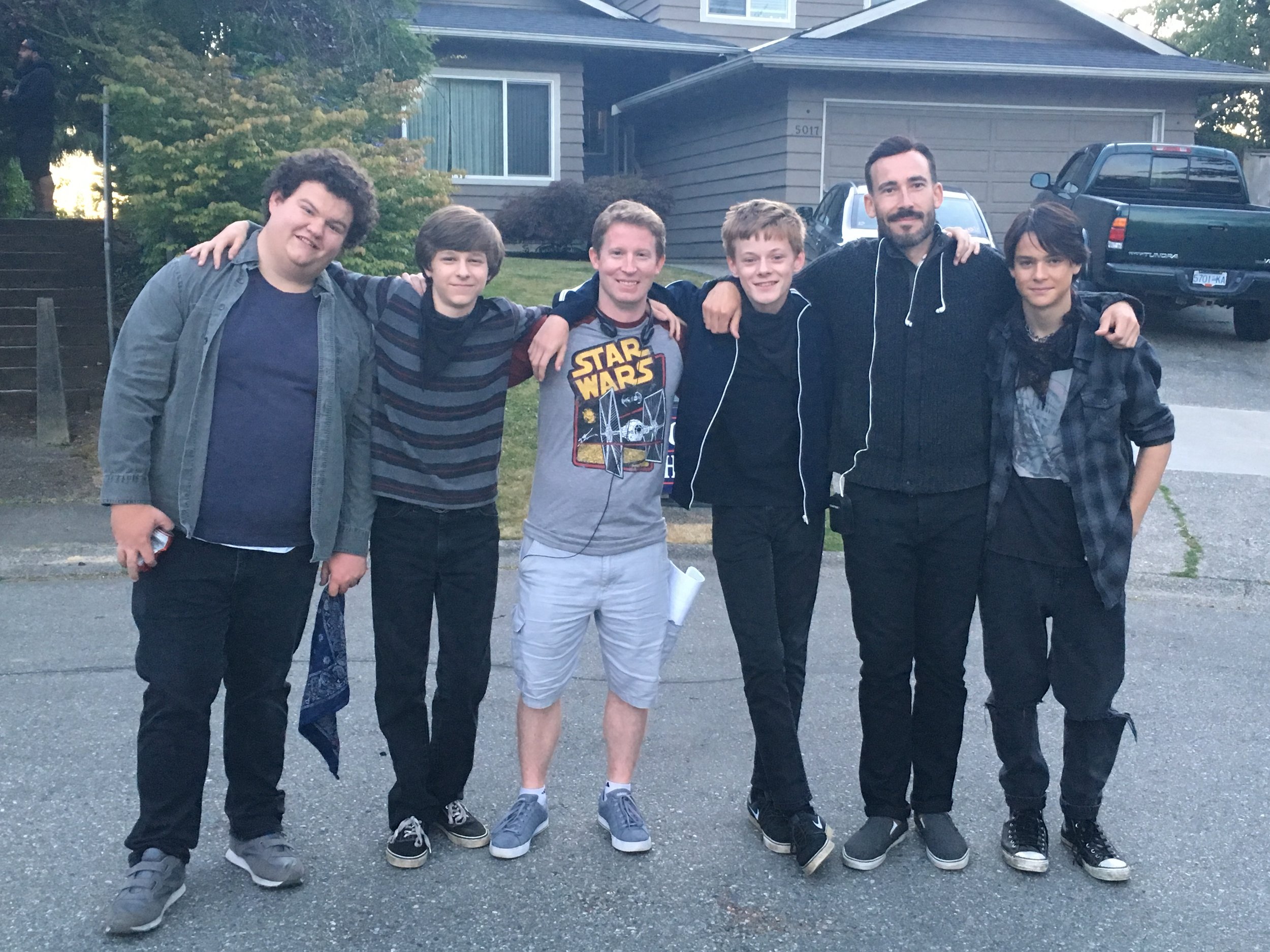 Group of six young people standing outside on a residential street, with arms around each other's shoulders, smiling at the camera.