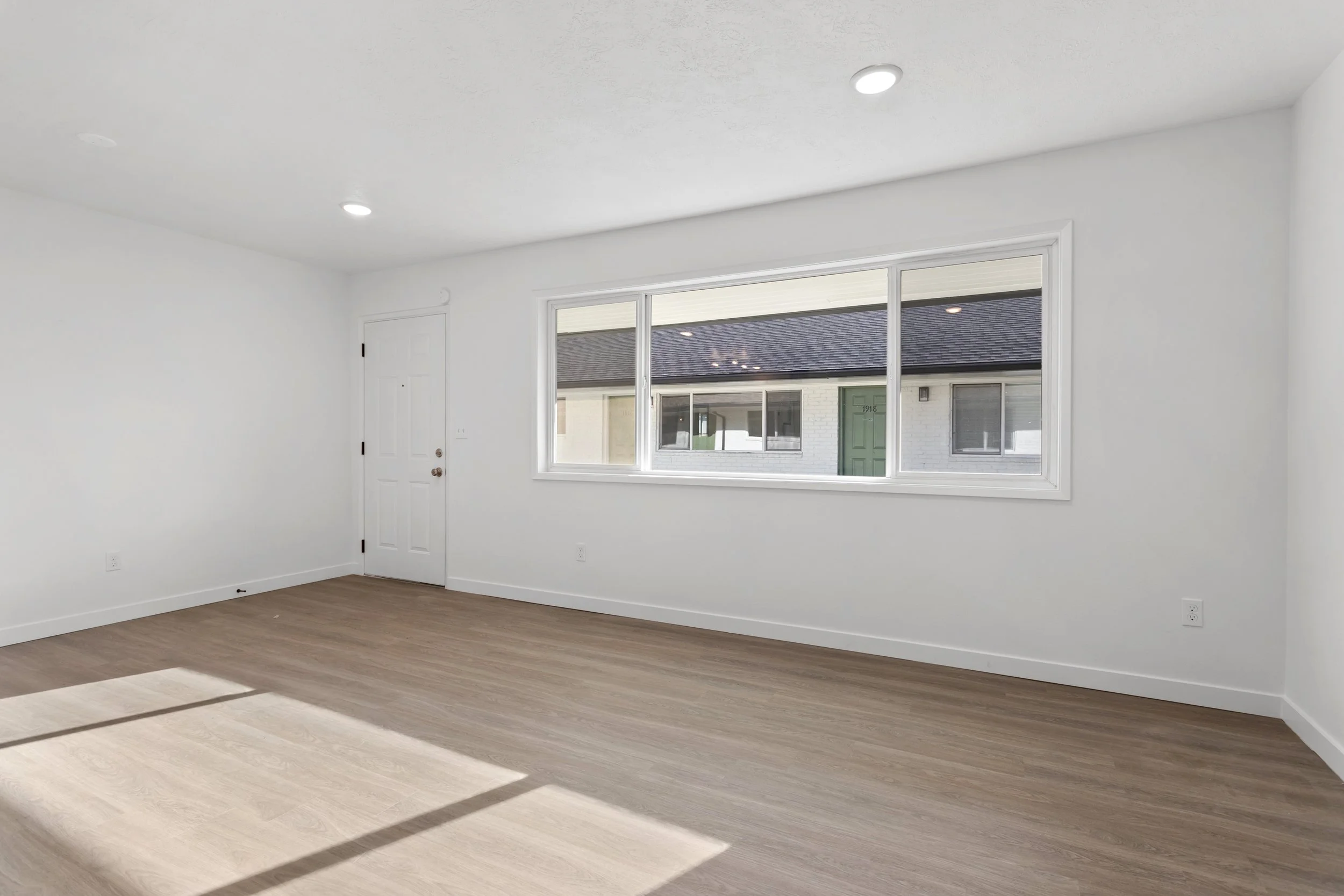 Empty room with large window, white walls, wooden floor, and a white door.