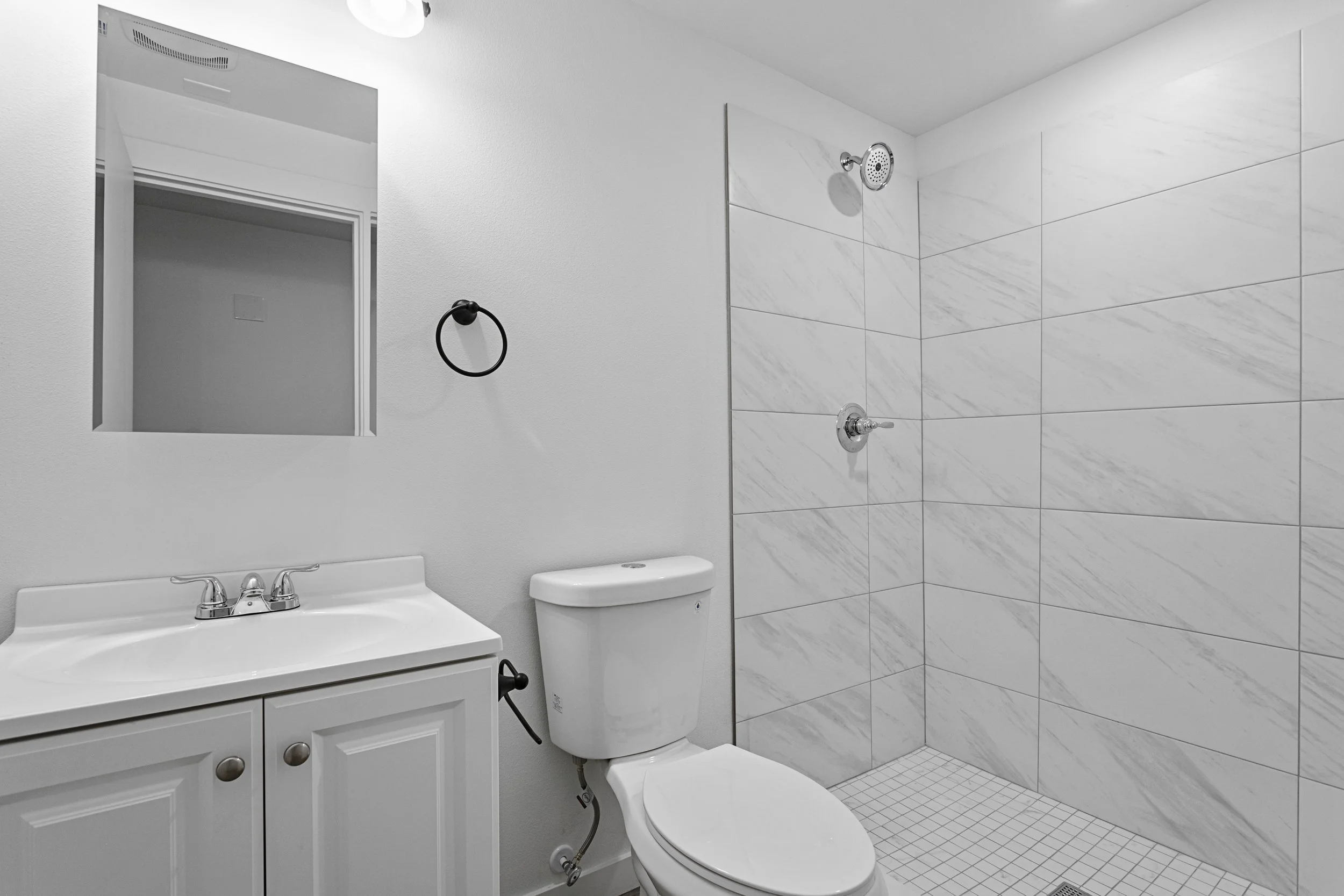 A clean, modern bathroom with a white vanity, mirror, toilet, and a walk-in shower with white marble tiles and a small square tile floor.