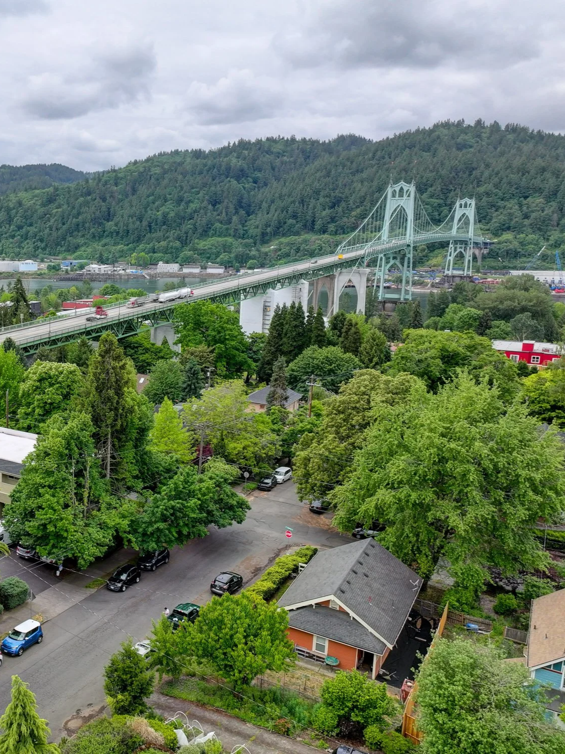 A suspension bridge over a river with a train crossing, surrounded by lush green trees and residential houses in a mountainous area under a cloudy sky.