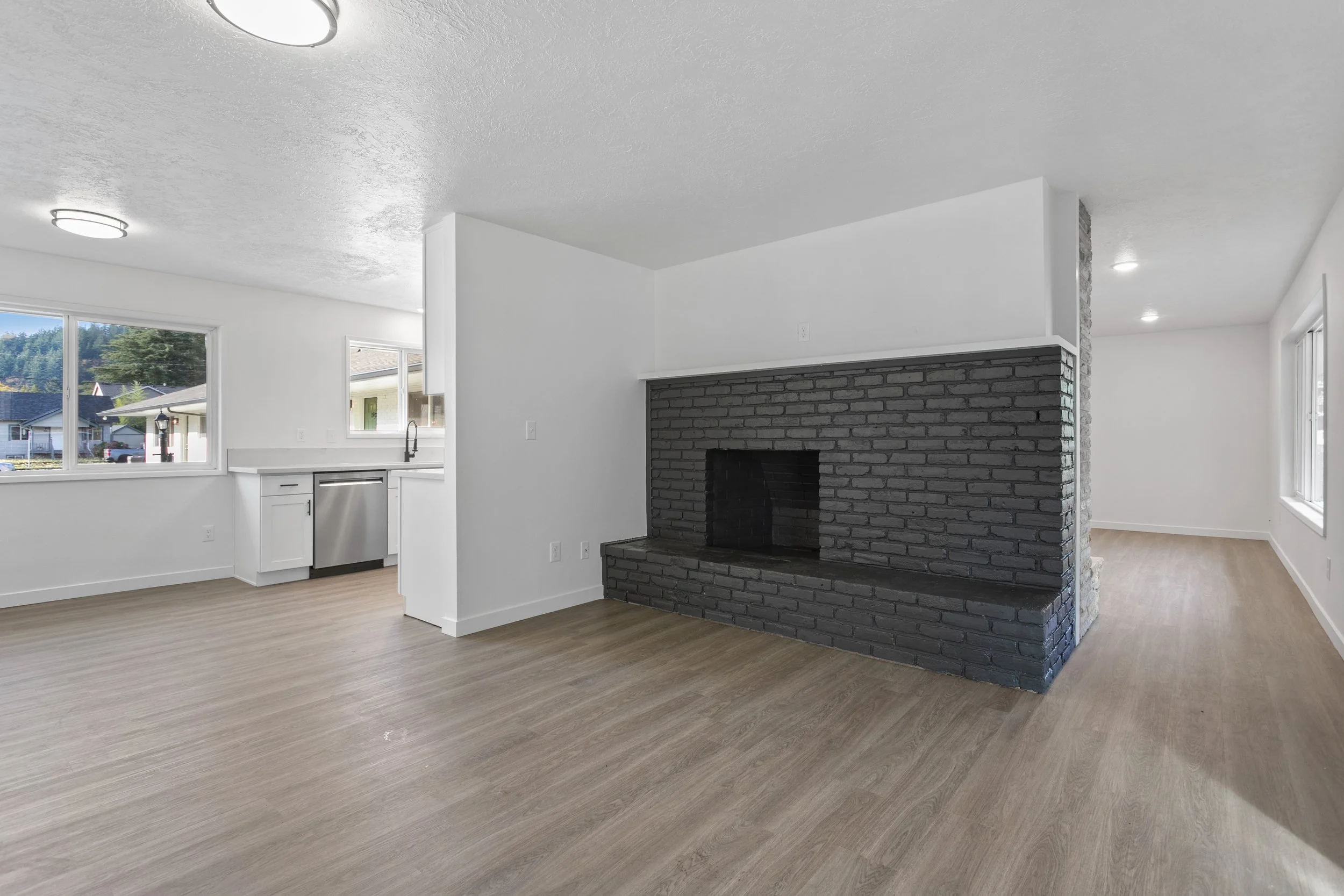 Empty living room with a dark brick fireplace, white walls, and light wood flooring, with large windows allowing natural light.
