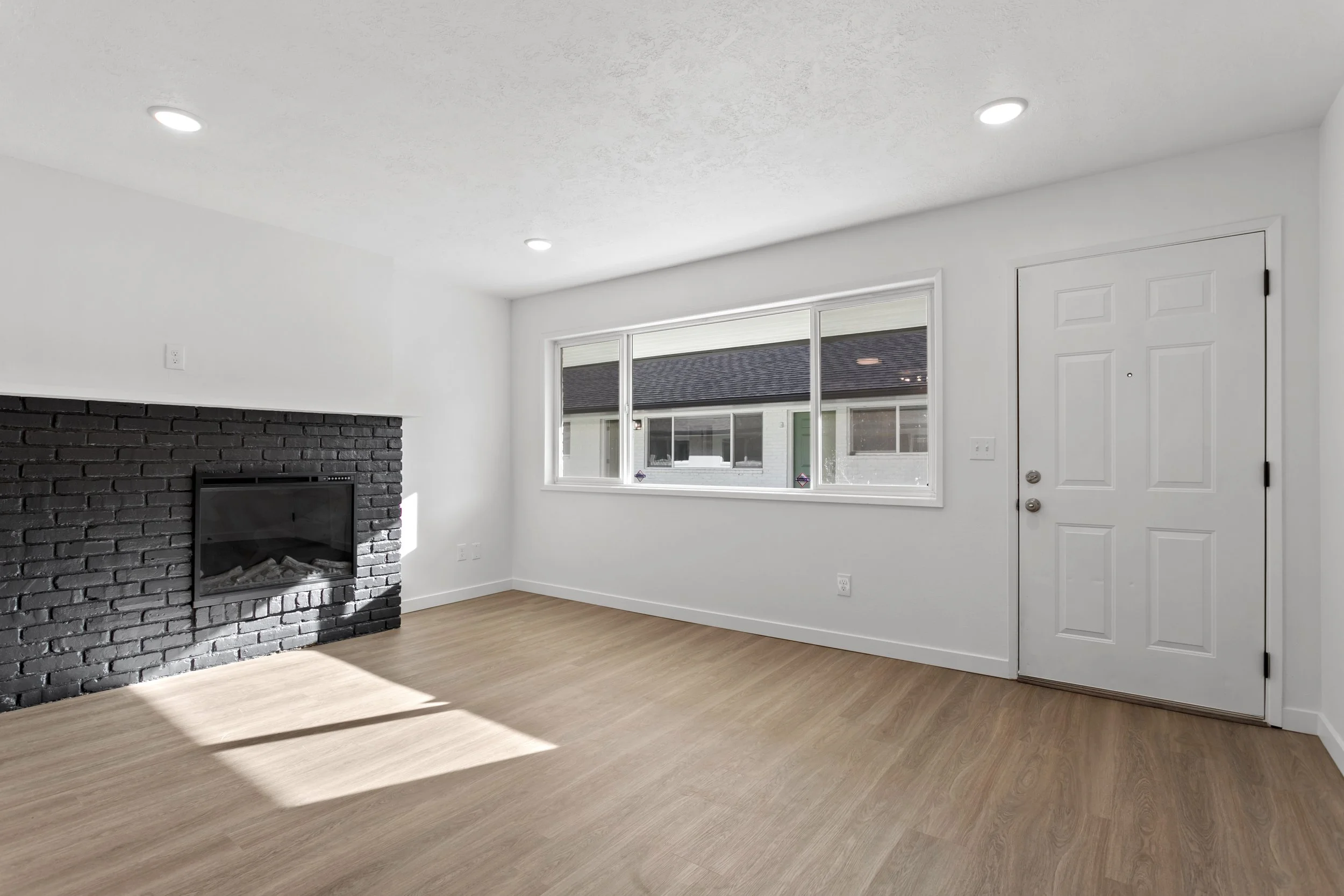 Empty living room with white walls, light wood floor, black brick fireplace, window facing outside, and a white door.