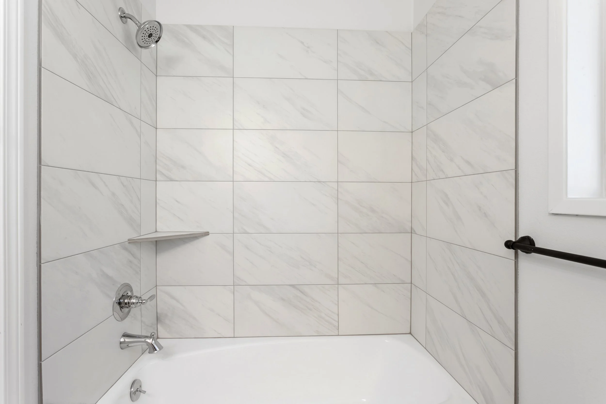 A clean shower with white marble tiled walls, a silver showerhead, and a black horizontal grab bar.