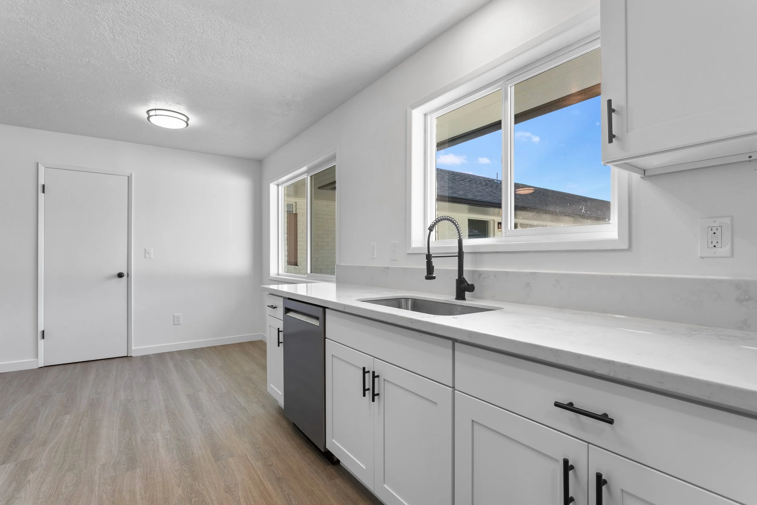 Modern kitchen with white cabinets, a black faucet, a marble countertop, and two large windows showing a blue sky and neighboring house.