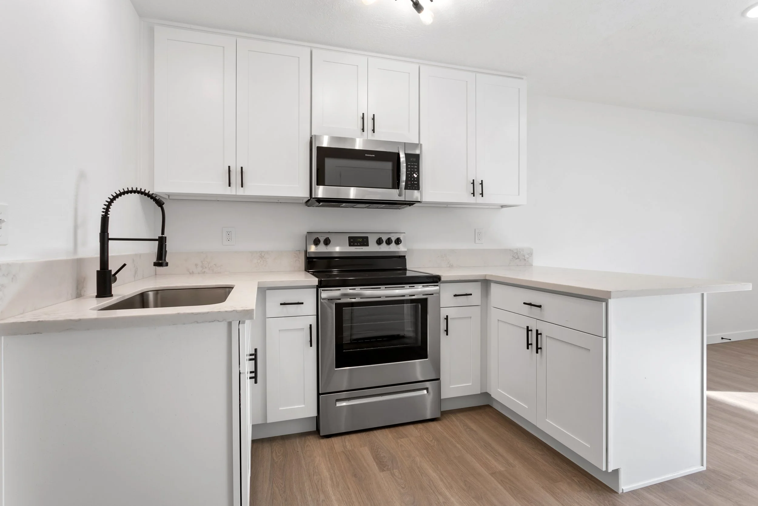 Modern kitchen with white cabinets, black handles, stainless steel microwave, oven, and a black faucet over a small sink, with light wood flooring.