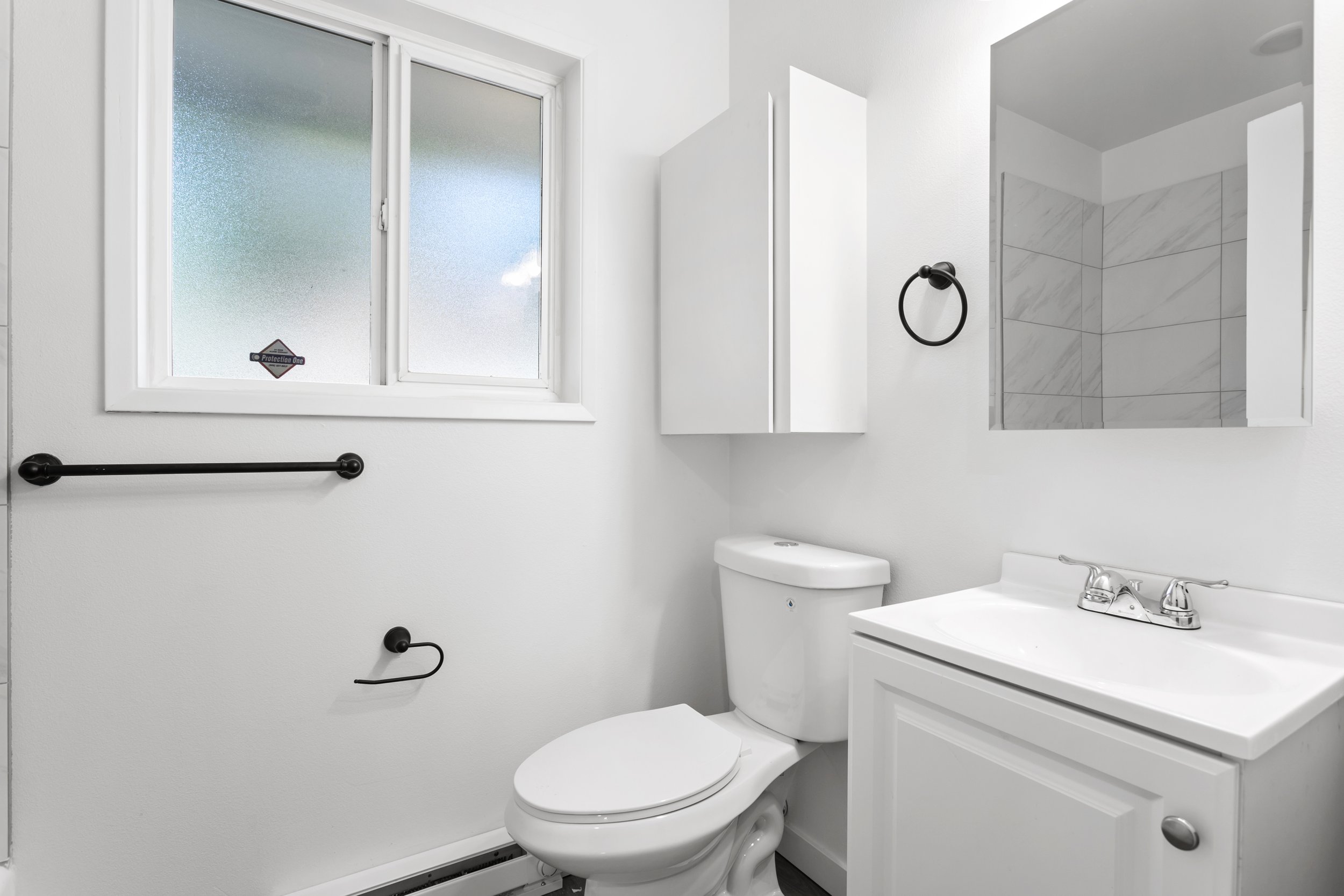 White bathroom with a frosted window, a toilet, a white cabinet, a black towel ring, a black toilet paper holder, a mirror above the sink, and a small mirror or medicine cabinet on the wall.