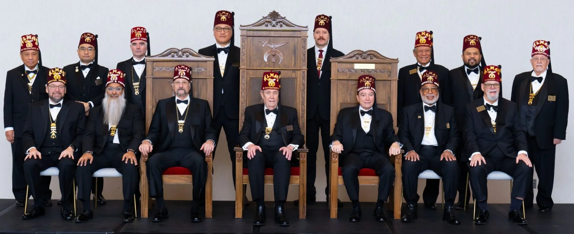 Group of 15 men in tuxedos and red fezzes with gold embroidery, sitting and standing in a formal setting. Some men are seated on chairs, others standing behind. The background features a plain light-colored wall and wooden thrones. The men are dressed in black tuxedos with bow ties, and some wear medals or badges.