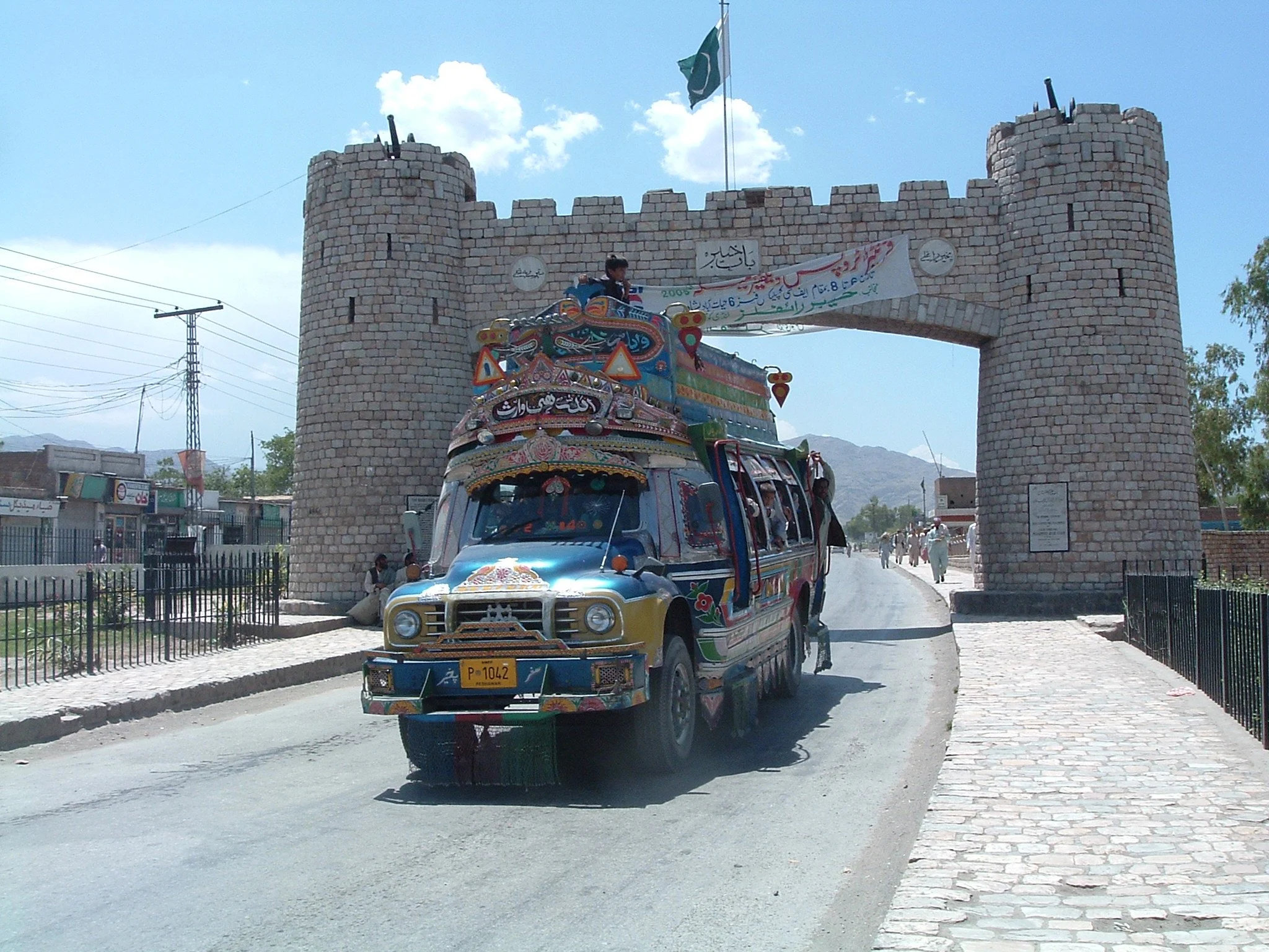 A colorful decorated truck passing through a historic stone gate with a flag flying above on a clear day, with mountains visible in the background.