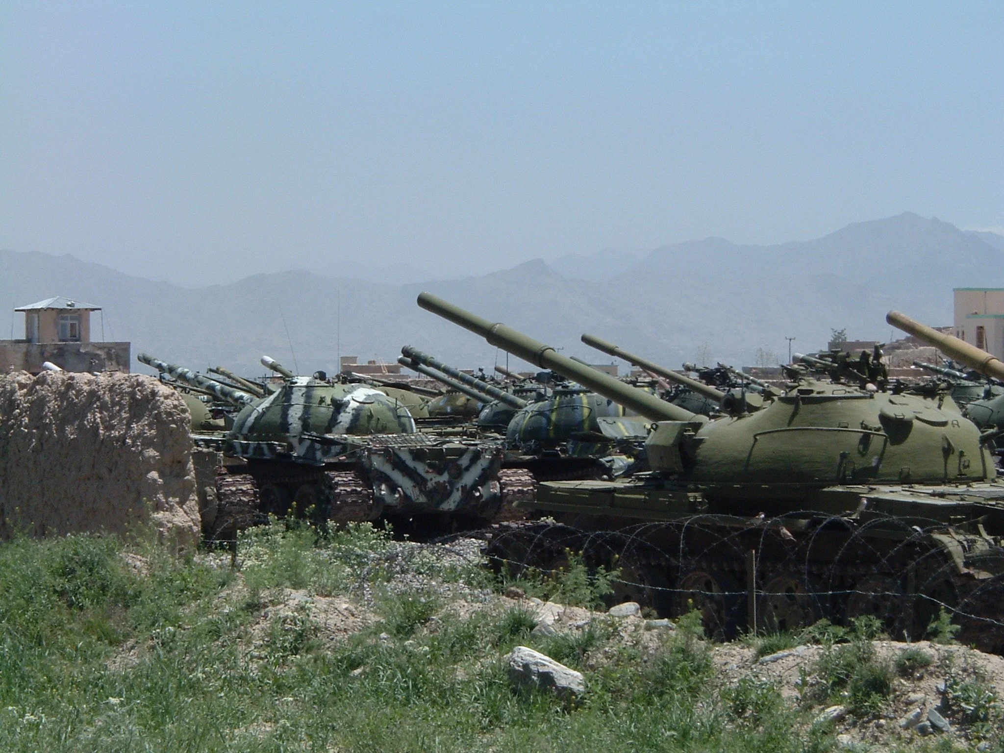 Multiple military tanks lined up outdoors with mountains in the background and barbed wire in the foreground.