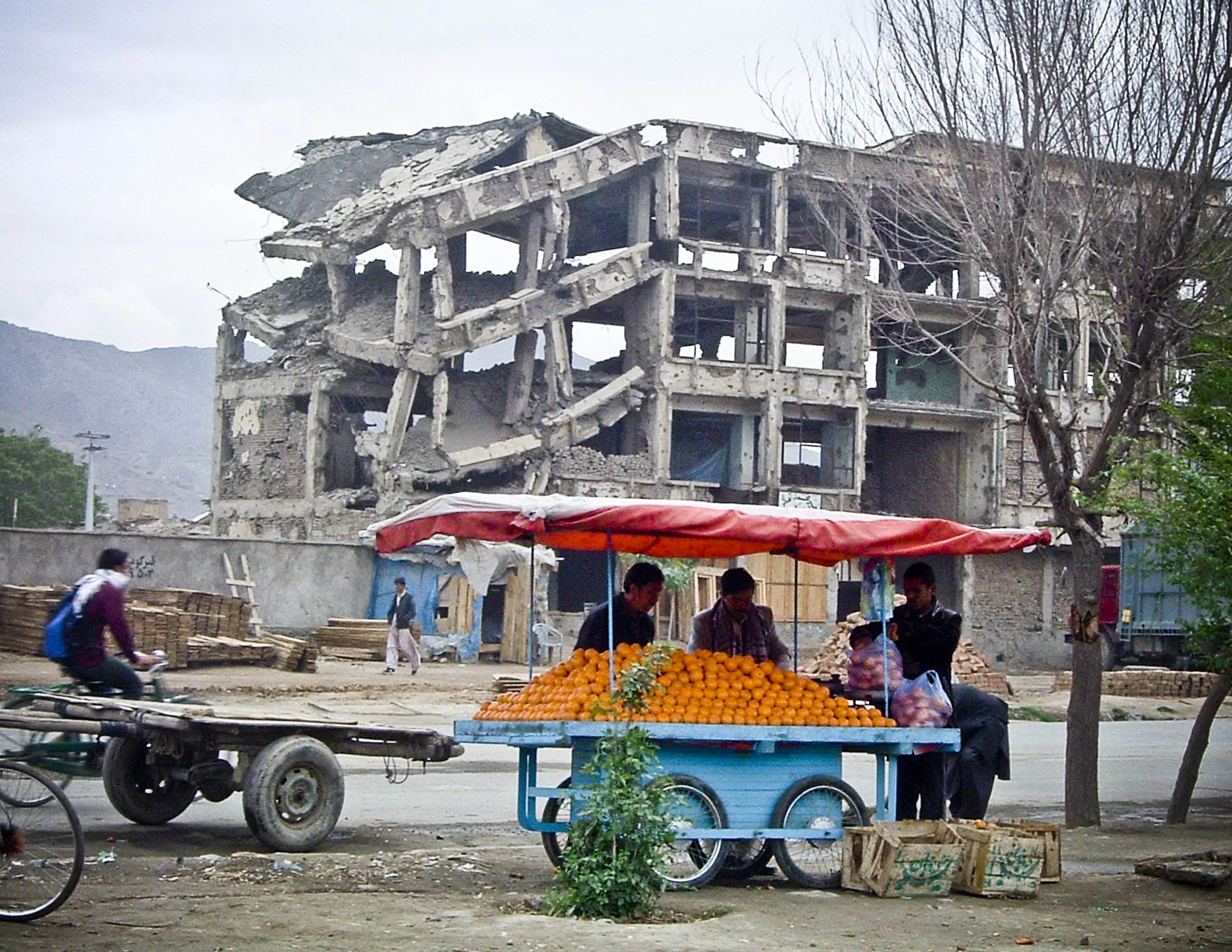 Street vendor selling oranges under a red canopy in front of a damaged building during daylight.