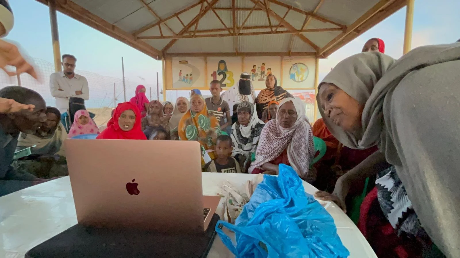 Group of people, including women and children, gathered around a table with a laptop, in an outdoor covered area with educational posters in the background.