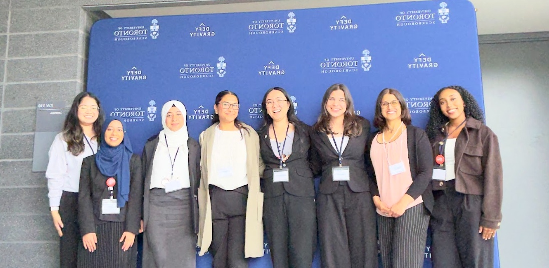 Group of eight women standing in front of a blue university of toronto backdrop, smiling at the camera.