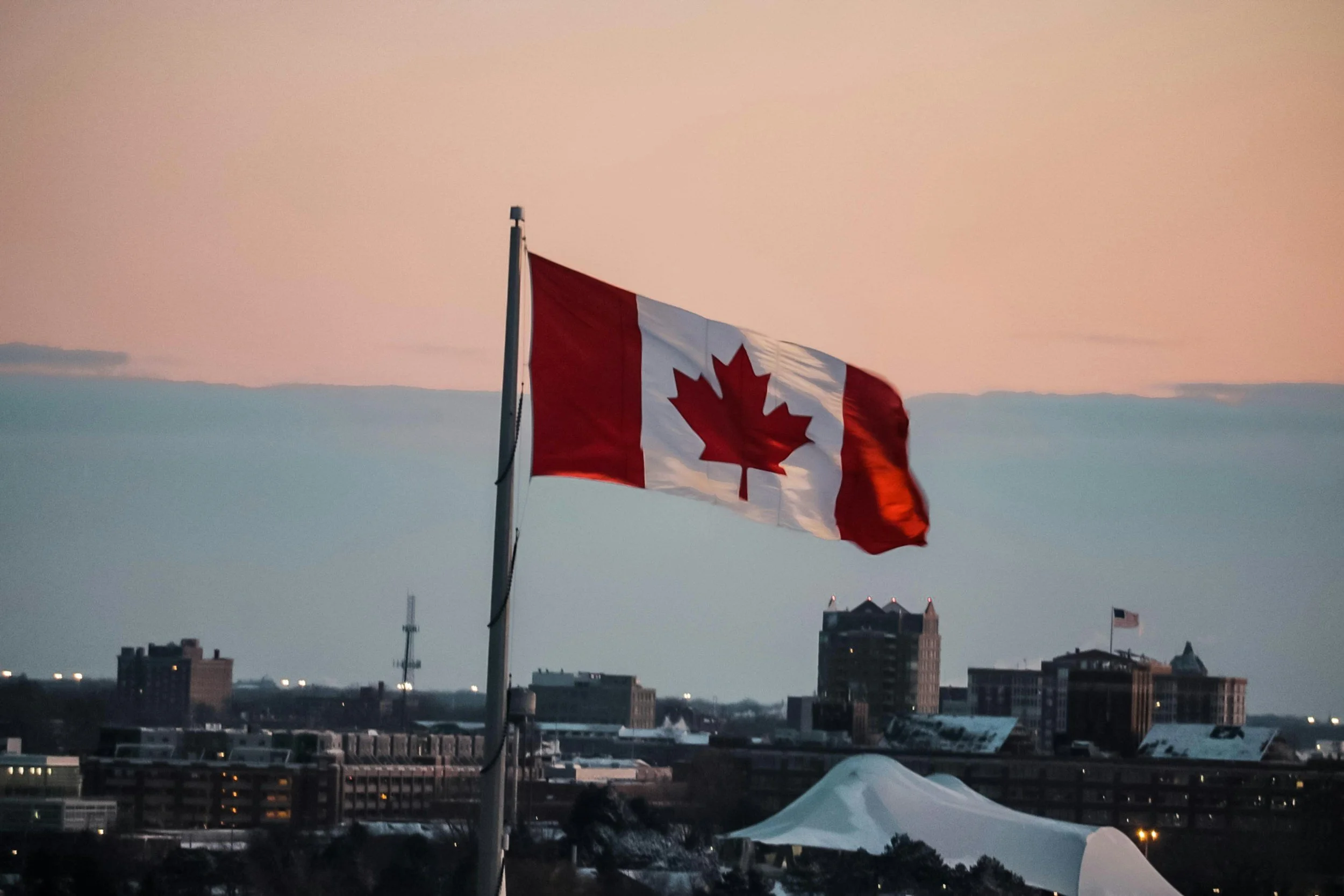 Canadian flag flying in front of a city skyline during sunset, with a pinkish sky and buildings in the background.