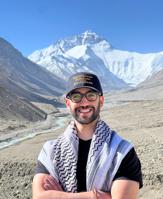 Man smiling in front of snow-capped mountains and a river in a rocky valley, wearing glasses, a black cap, and a keffiyeh scarf.