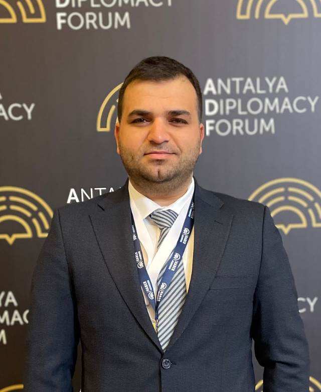 A man in a suit and striped tie standing in front of a backdrop with the text 'Antalya Diplomacy Forum' and a logo.