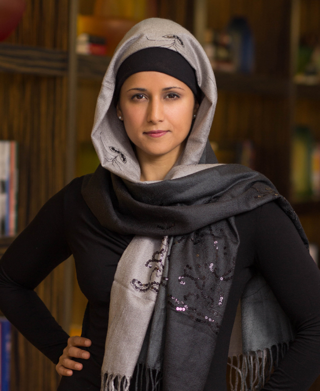 A woman wearing a black shirt and a gray scarf with sequins and embroidered details, standing with her hand on her hip in front of a bookshelf.