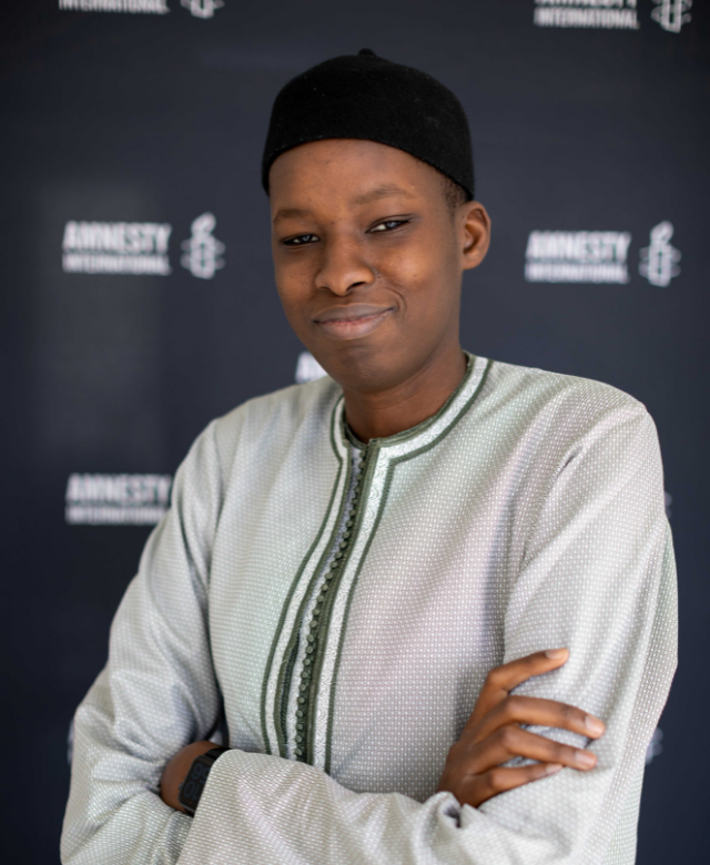 A young person with short hair wearing a black head covering and a light-colored traditional garment, standing against a backdrop with 'Amnesty International' logos, looking confidently at the camera with arms crossed.