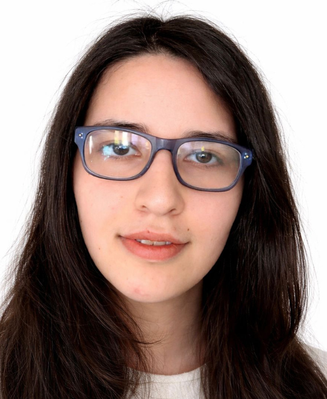 Close-up portrait of a young woman with long dark hair, wearing glasses and a white top, smiling subtly against a white background.
