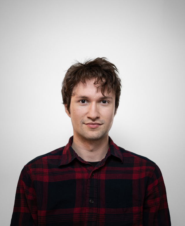 A young man with light skin, brown hair, and a slight smile, wearing a red and black plaid shirt, standing against a plain white background.