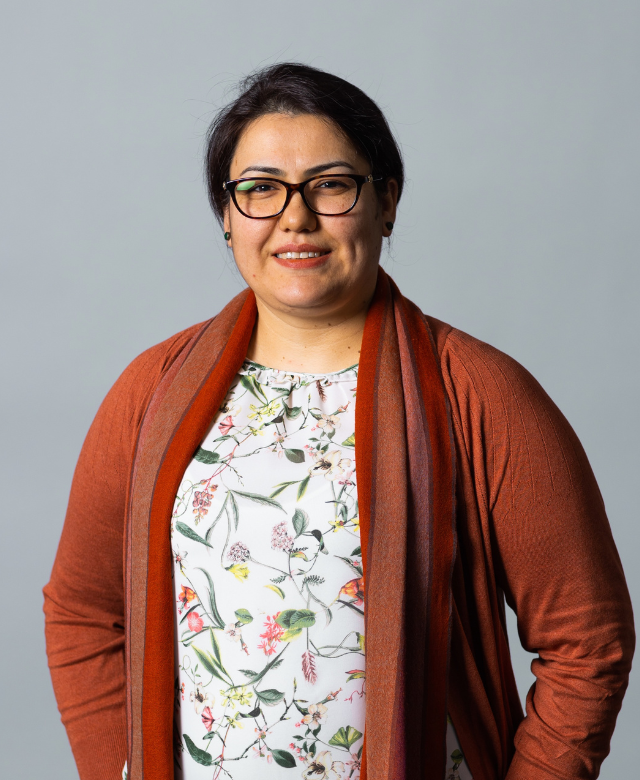 A woman with glasses wearing a floral blouse and an orange-brown cardigan, smiling and standing against a plain grey background.