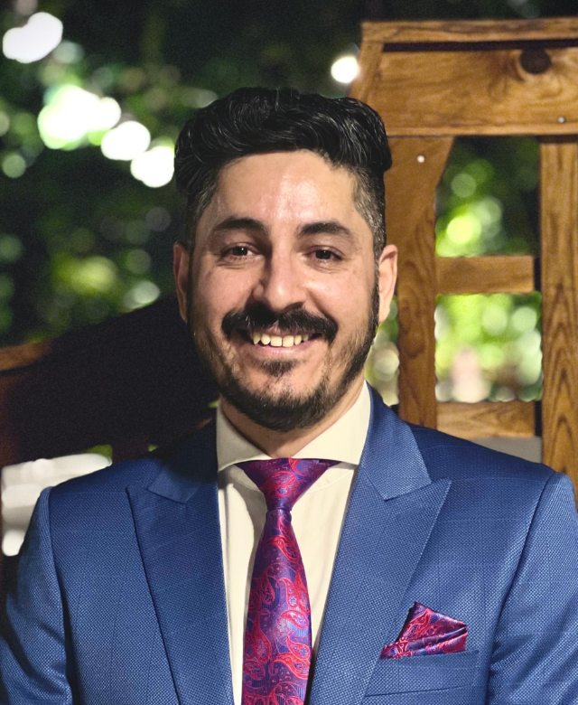 A man with dark hair and a beard, dressed in a blue suit, white shirt, and paisley tie, smiling at the camera outdoors with blurred lights and greenery in the background.