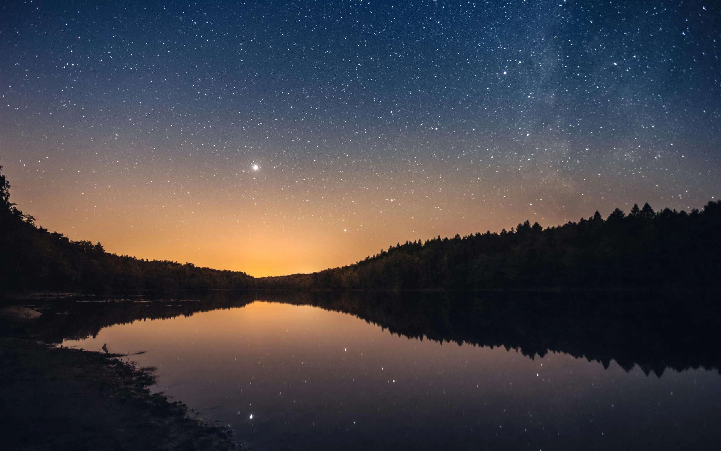Nighttime landscape with a star-filled sky, a faint orange glow on the horizon, and a calm river reflecting the sky and trees