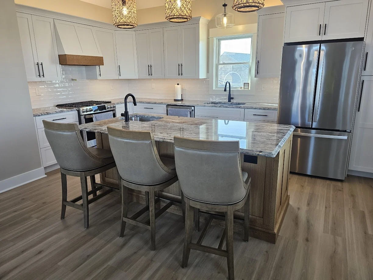 Modern kitchen with white cabinets, stainless steel refrigerator, granite countertops, and a wooden kitchen island with seating for three, hardwood floors, pendant lights, and a window above the sink.