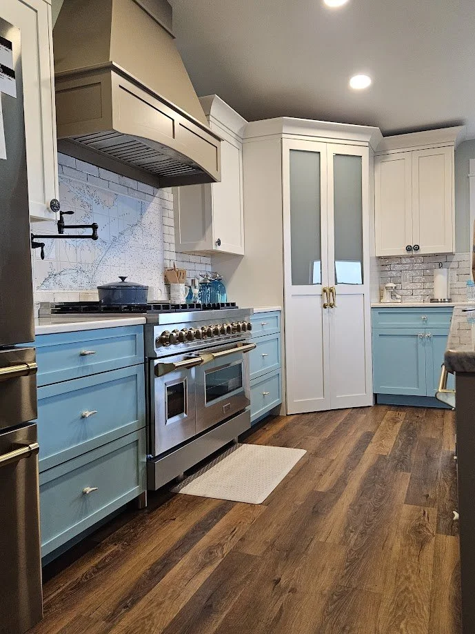 Modern kitchen with light blue and white cabinets, stainless steel oven and stovetop, wooden flooring, and decorative backsplash.