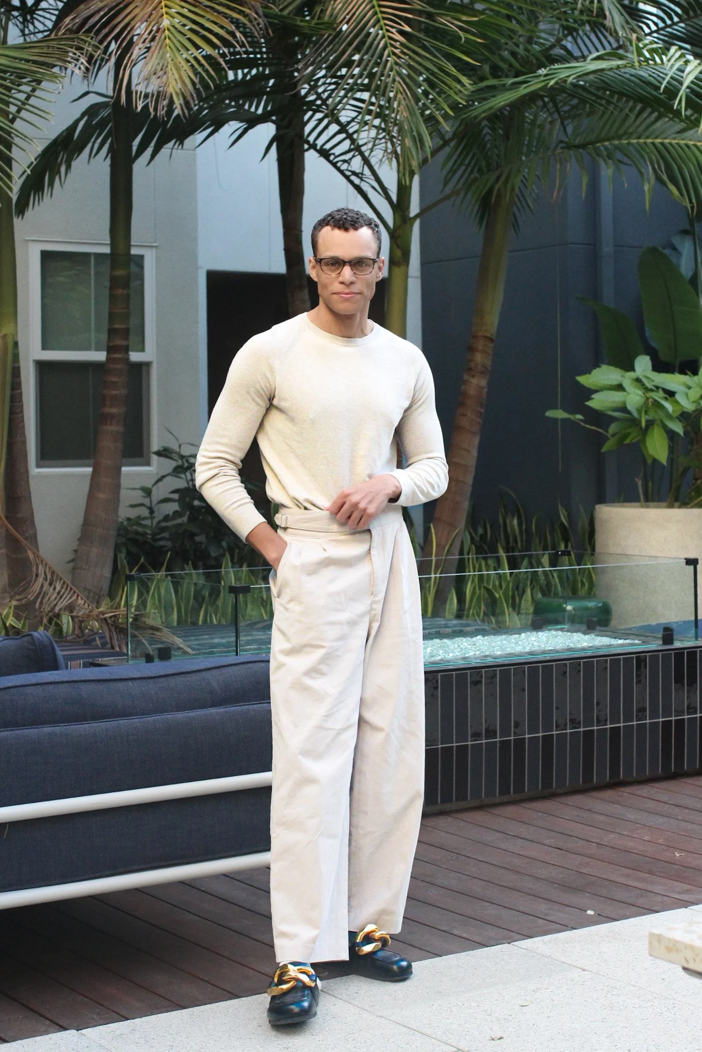 A man wearing glasses, a beige sweater, light-colored high-waisted baggy pants, and black shoes with gold chain details standing outdoors near a patio with tropical plants and modern buildings in the background.