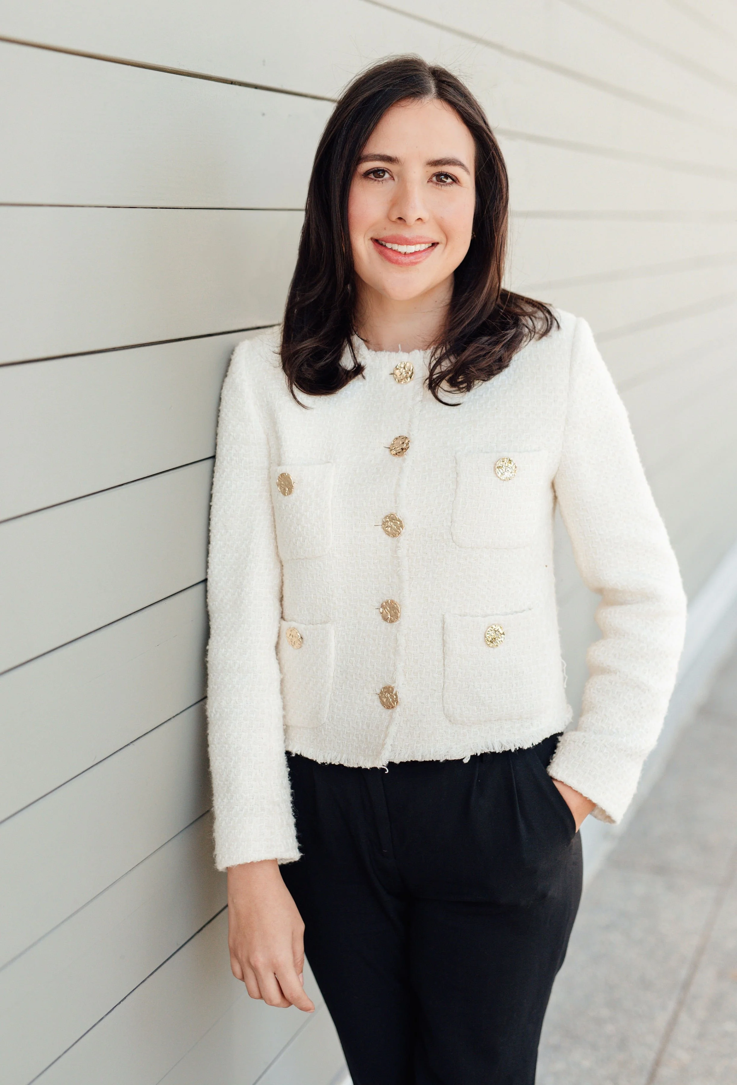A woman with dark brown hair wearing a cream-colored jacket with gold buttons, smiling and standing against a light gray wooden wall.