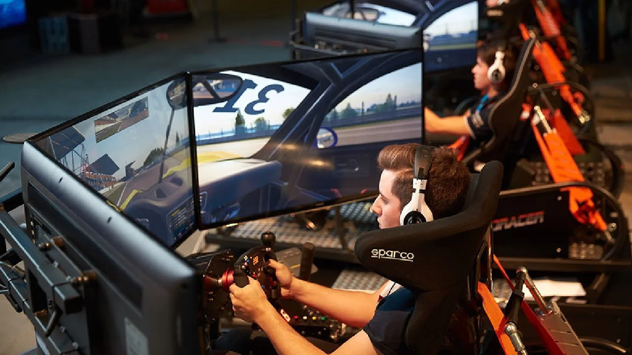 Two people in racing simulators, wearing headphones, playing a driving video game on curved monitors.